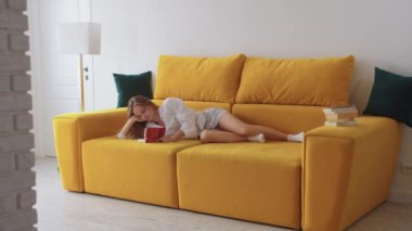 Sarı kanepede uzanmış kitap okuyan genç bir kadın. Orta çekim
