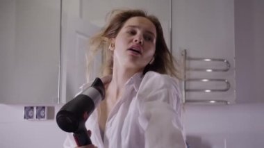 Genç bir kadın uzun saçlarını aynanın önünde saç kurutma makinesiyle kurutur ve şarkı söyler. Kamera Manzara Noktasıdır. Portre