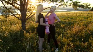 Gün batımında elinde yoga minderleriyle sahada duran iki genç sarışın kadın. Orta çekim