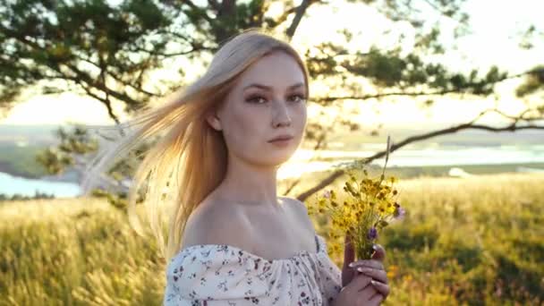 一个金发碧眼的年轻女人拿着花 看着站在田野里的摄像机 肖像画 — 图库视频影像