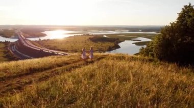 Yaz ışığı elbiseli iki genç sarışın kadın el ele tutuşup tepeden aşağı koşuyorlar. Hava görünümü