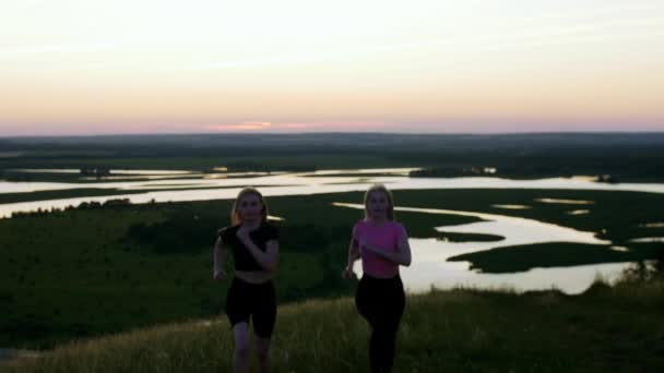 2人の若いスポーツブロンドの女性が夕日のフィールドでジョギングし カメラを見ています ミッドショット — ストック動画