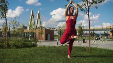 Kırmızı eşofmanlı bir kadın dışarıda yoga yapıyor. Tek ayak üstünde duruyor. Orta çekim