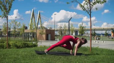 Kırmızı eşofmanlı yetişkin bir kadın köprünün yanındaki parkta spor egzersizleri yapıyor. Orta çekim