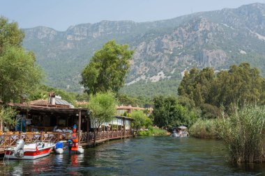 Akyaka, Mugla, Turkey - July 30, 2016. Azmak stream in Akyaka village in Mugla province of Turkey, with restaurant, boats and Sakar mountains in the background.