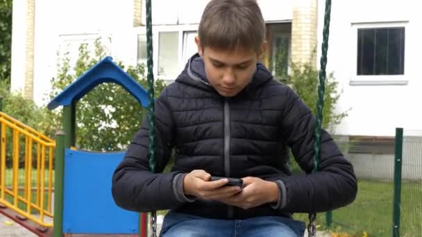 Kaukasischer Junge sitzt auf Schaukel auf Spielplatz im Freien und benutzt sein Handy, spielt Videospiel, surft im Internet, sendet SMS — Stockvideo