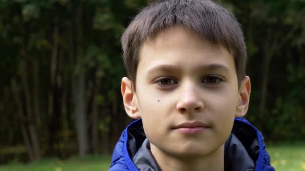 Close up portrait of teenage boy in blue jacket outdoors with city park trees in background. 4K video — Stock Video