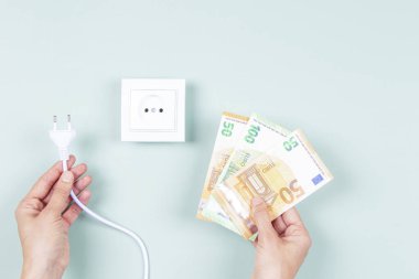 Elektrik maliyeti ve pahalı enerji konsepti. Elektrik prizi ve Avro para banknotlarını açık yeşil arka planda beyaz elektrik soketinin yanında tutan kadın eller