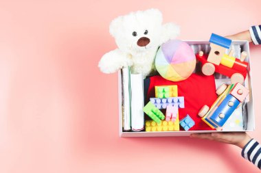 Bağış konsepti. Çocuk elleri, pembe arka planda giysiler, kitaplar ve oyuncaklarla dolu bağış kutusunu tutuyor. Üst görünüm