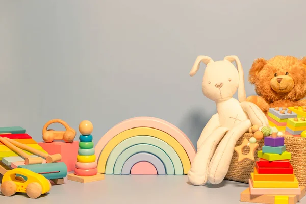 Toys background. Toy box with teddy bear, bunny and wood rattle. Educational wooden Montessori toys on pastel blue background. Cute toys collection for small children. Front view