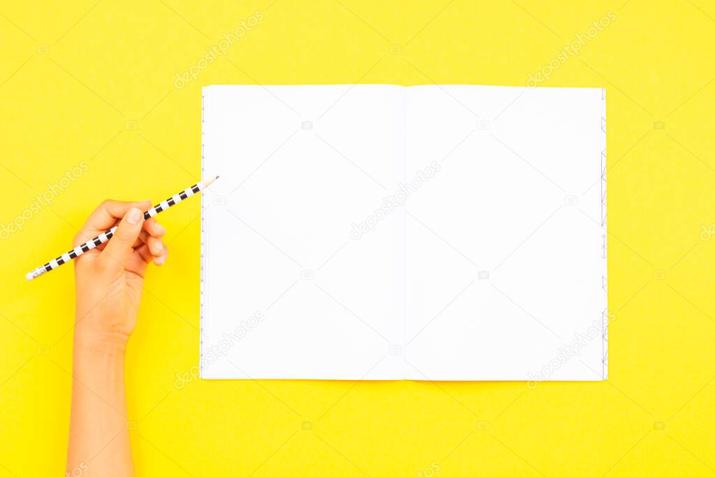 Left hand holding pencil for writing on blank open notebook over yellow background. Top view, copy space for text. Left Handers Day