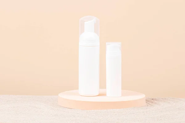 Two white blank plastic packaging cosmetic pump bottles for mousse foam cream and lotion stand on round podium, beach sand and beige background. Front view