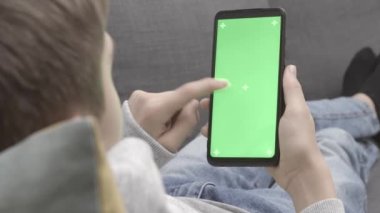 Arka planda, yeşil ekran krom arka planda akıllı telefon kullanan bir genç oturuyor. Sosyal ağlar konsepti. Video izlemek, kaydırmak, ekrana dokunmak