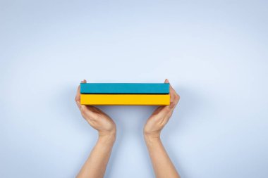 Açık mavi arkaplan üzerinde iki mavi ve sarı renkli kitabı tutan kadın eller. Eğitim, kendi kendini öğrenme, kitap değişimi