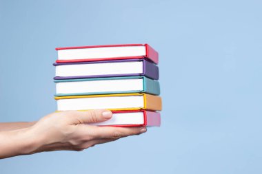 Açık mavi arkaplan üzerinde kitap yığını tutan kadın eller. Eğitim, kendi kendine öğrenme, kitap değiştirme, hobi, dinlenme zamanı.