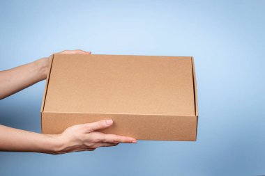 Kadın elleri, doğal kıvrımlı kartondan yapılmış kahverengi ekolojik paket kutusunu tutuyor. Mavi arkaplandaki posta kutusunun maketi. Paketleme, alışveriş, teslimat konsepti