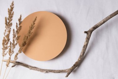 Boş yuvarlak bej platform podyumu, kuru ağaç dalları ve beyaz keten kumaş arka planda eğilmiş çimenler. Kozmetik veya ürün sunumu için en az yaratıcı kompozisyon. Üst görünüm