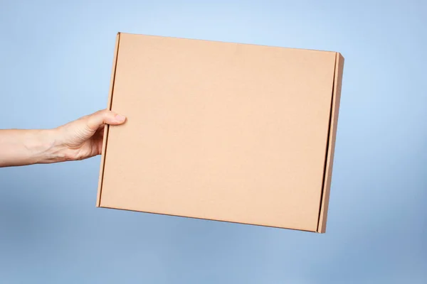Mulher mão segurando caixa de pacote ecológico marrom feito de papelão ondulado natural. Mockup caixa de envio de encomendas no fundo azul. Embalagem, compras, conceito de entrega — Fotografia de Stock