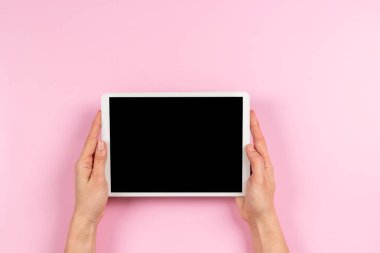 Woman hand holding digital tablet computer with blank screen mock up on pastel pink color background. Top view, copy space.
