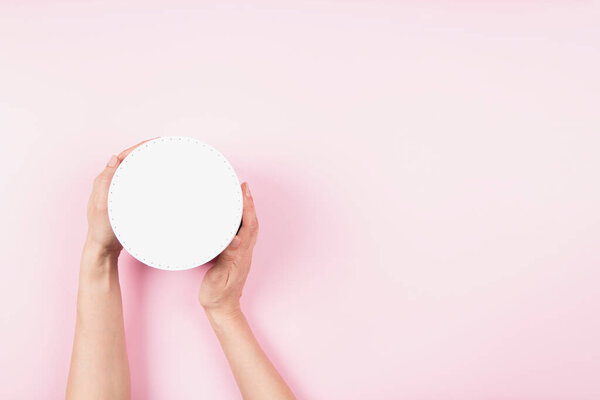 Woman hands holding white round cardboard box with cap onpastel pink background. Mock up package box. Top view