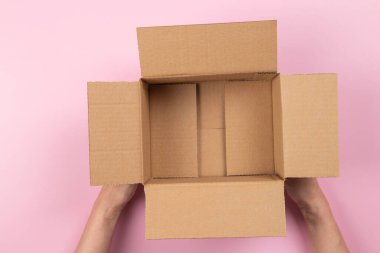 Female hands with empty open brown cardboard box on pastel pink background. Top view