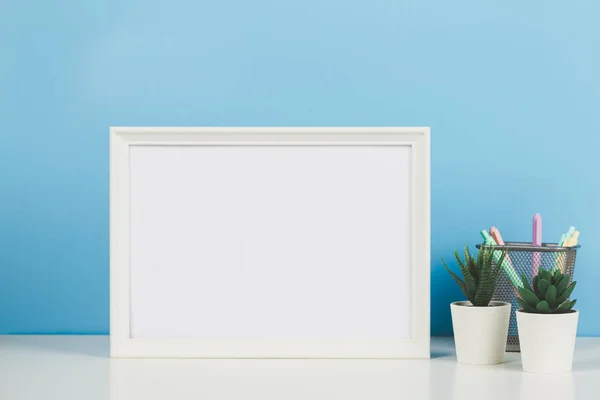 White empty wooden mock up frame and succulent flowers on white desk with light blue background. Front view.