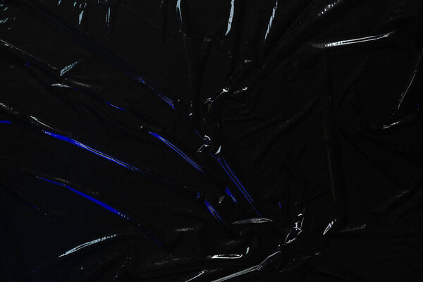 Transparent plastic wrap on black background. Crumpled wrinkled plastic cellophane. Reflecting blue color light and shadow on creases and folds in plastic surface. Texture overlay effect template.