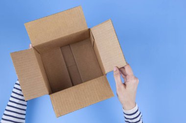 Female hands holding open empty brown cardboard box on light blue background. Top view