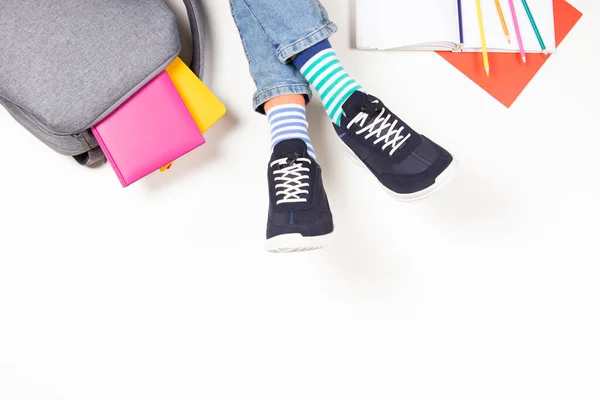 Kid wearing different pair of socks. Child foots in mismatched socks and colorful sneakers, backpack with books and school supplies on white background. Top view, copy space