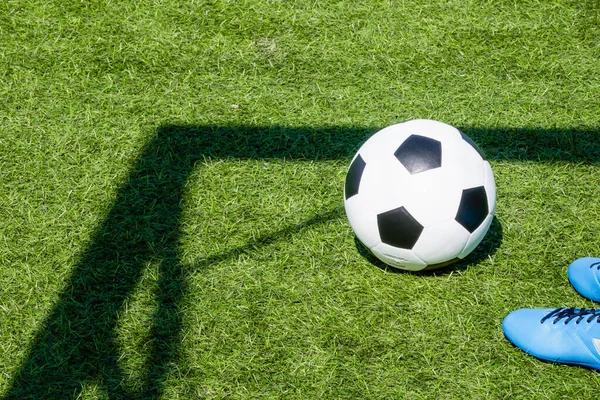 Green artificial turf soccer field with shadow from football goal net and soccer ball on sunny day outdoors. Football soccer sport background.