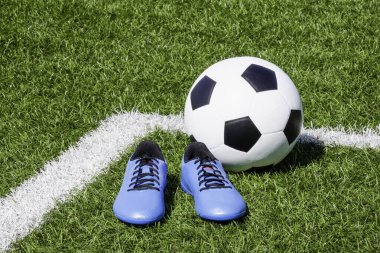 Soccer ball and pair of soccer football sports shoes cleats on green artificial turf football field with white lines.