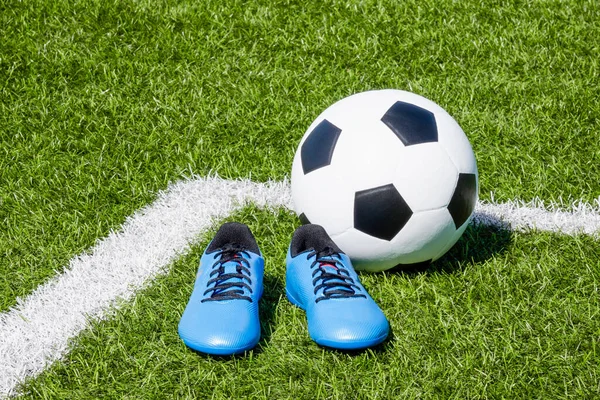 Soccer ball and pair of soccer football sports shoes cleats on green artificial turf football field with white lines.