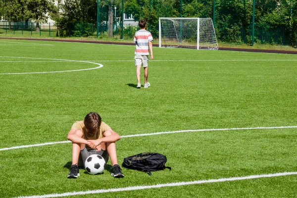 Sad disappointed teenage boy sitting in empty school sport stadium outdoors. Friend walks away and leaves his friend sitting alone. Emotions, defeat, lost game, difficulties, problems of teenagers.
