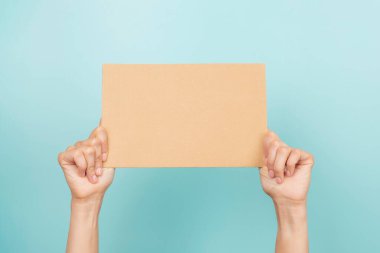 Woman hands holding blank sheet of brown cardboard on light blue background.