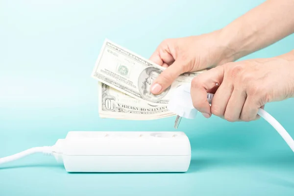 Woman hands holding electric power plug and US dollar banknotes near power strip extension cord on blue background. Electricity cost, electric prices increase, expensive energy concept.