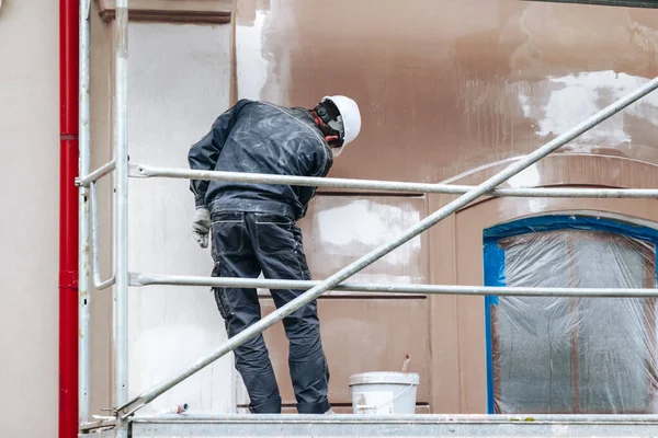 Renovation, restoration, refurbishment. Unrecognizable worker renovating wall of classical style building, standing on scaffolding. Construction worker prepares house facade wall for painting outdoors