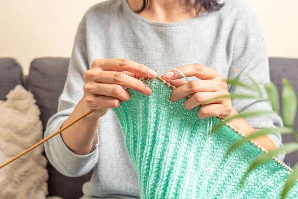 Female hands holding bamboo and knitting needles knitting green woolen sweater. Middle aged woman sitting on sofa at home and knitting. Hobby, relaxation, mental health, sustainable lifestyle.