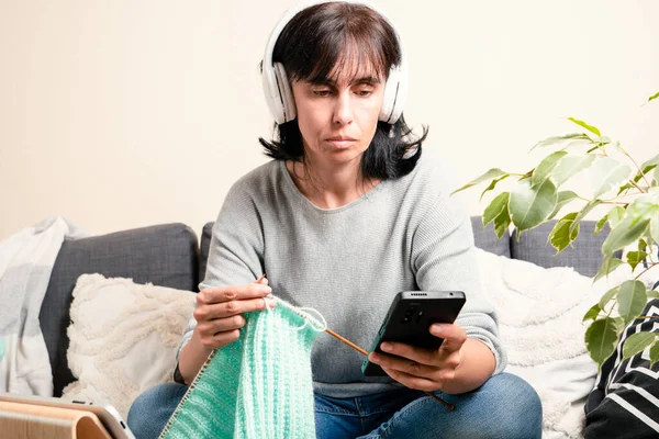 Middle age woman at home look at digital tablet computer and knitting. Woman learning, watching video tutorial, online knitting class lesson, follow knitting patterns, listen audio book,learn language