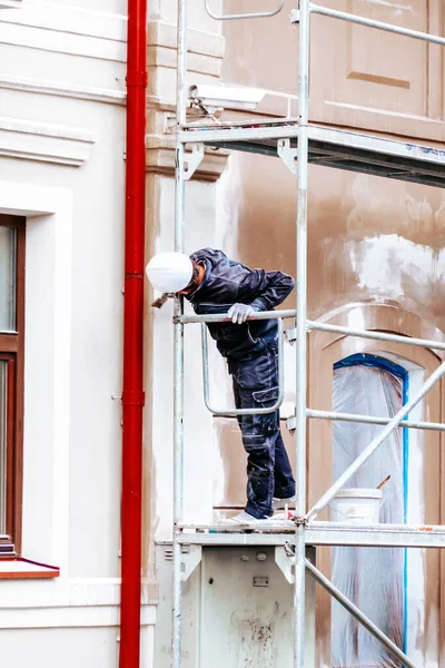 Renovation, restoration, refurbishment. Unrecognizable worker renovating wall of classical style building, standing on scaffolding. Construction worker prepares house facade wall for painting outdoors