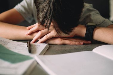 Hayal kırıklığına uğramış genç çocuk kafasını eğip kitap yığınları, ders kitapları, alıştırma kitapları arasında oturuyor. Çevrimiçi öğrenme, okuma güçlüğü, eğitim kavramı.