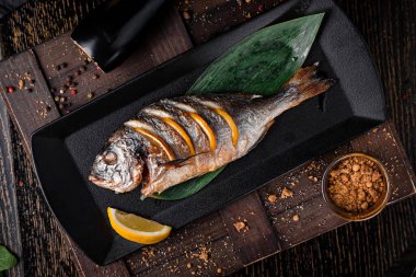 Fırında kızartılmış Dorado balığı ve ahşap bir masada siyah bir tabakta limon dilimleri.