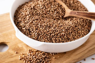 Kuru karabuğday, tahta bir tahtanın üzerinde beyaz bir kasede, tahta bir kaşıkla, beyaz arka planda, üst tarafta, yakın planda inliyor.