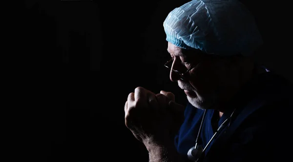 An elderly doctor with a stethoscope and a cap on a black background in profile, holds his hands near his face, prays, close-up, space for text, banner