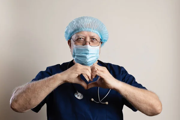 An elderly doctor with a stethoscope, a cap, a mask and glasses on a light background, looks kindly into the camera, shows a heart symbol with a gesture, close-up, space for text