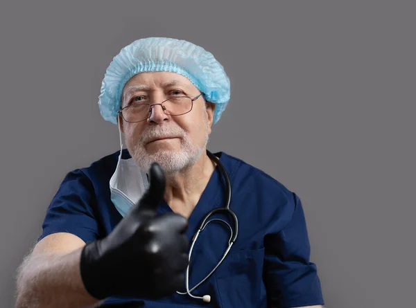 An elderly doctor with a stethoscope, a cap, a mask and glasses on a gray background, looks kindly at the camera, shows a like gesture, close-up, space for text