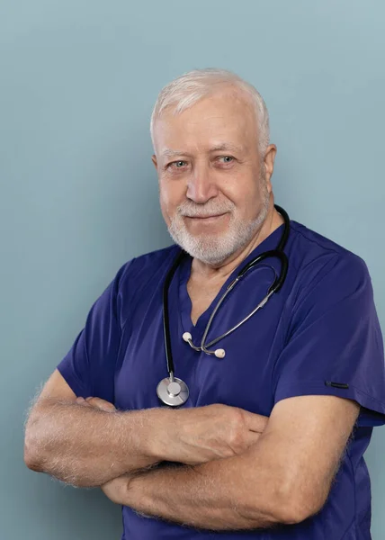 An elderly gray-haired doctor with a stethoscope stands with his arms folded on his chest on a light background, looks to the side, portrait, close-up