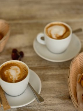 Fotoğrafta iki fincan kahveli hamur işleri var. Kahvaltı ahşap bir masanın üzerindeki bir kafede yer alır. Arka planda tropikal bitki örtüsü görünür..
