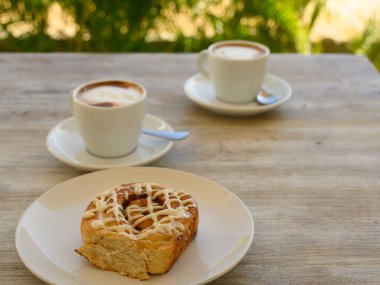 Fotoğrafta iki fincan kahveli hamur işleri var. Kahvaltı ahşap bir masanın üzerindeki bir kafede yer alır. Arka planda tropikal bitki örtüsü görünür..