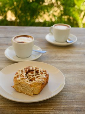 Fotoğrafta hamur işleri ve bir fincan kahve var. Kahvaltı ahşap bir masanın üzerindeki bir kafede yer alır. Arka planda tropikal bitki örtüsü görünür..