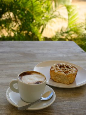 Fotoğrafta iki fincan kahveli hamur işleri var. Kahvaltı ahşap bir masanın üzerindeki bir kafede yer alır. Arka planda tropikal bitki örtüsü görünür..